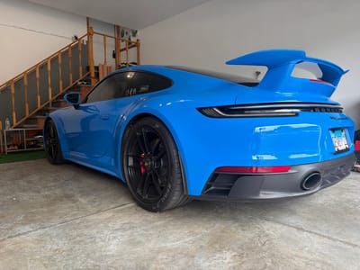 In a garage with a concrete floor, a bright blue Porsche 911 Carrera GTS is parked. The car is viewed from the rear-left quarter angle. It features a large rear spoiler and black wheels with red brake calipers. A license plate from Illinois is visible on the back. To the left of the car, a wooden staircase with a dark railing leads upstairs. Various items are stored on shelves below the stairs, including shoes and bottles. The walls of the garage are white, and there are some decorative plants on the wall above the stairs.