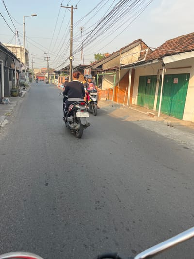 The image depicts a street scene in Sugih Waras, Indonesia, during the late afternoon. The foreground shows the handlebars of a motorcycle, indicating the image was taken from a moving vehicle. The road is paved and asphalt. In the midground, two people are riding on a motorcycle, with the driver wearing a black shirt and patterned pants, and a passenger behind, wearing a pink headscarf and holding a small child. Another motorcycle is visible further ahead, with two occupants. The street is lined with buildings, some appearing to be shops or small residences, with tiled roofs and green shuttered doors. Electrical wires are strung overhead on utility poles. The lighting suggests a clear sky with the sun casting shadows.