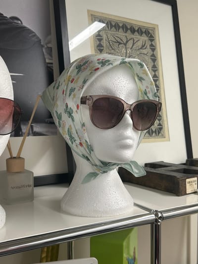 The image displays an indoor scene in Garston, United Kingdom, focused on a white styrofoam mannequin head positioned centrally on a white shelf. This mannequin wears a light green headscarf patterned with red strawberries and green leaves, tied beneath its chin, and light brown-framed, dark-tinted sunglasses. To the immediate left, a portion of a second white styrofoam mannequin head is visible, adorned with red-tinted sunglasses. Adjacent to the partial mannequin head is a frosted glass diffuser bottle, labeled "AromaWorks," featuring a wooden cap and multiple reeds. The background includes two framed artworks: a tall, black-framed monochromatic print on the left, and a smaller, black-framed print with intricate dark patterns on a light background to the right. A dark, multi-chambered wooden object with a white sticker is situated on the far right of the shelf. The white shelf surface is supported by a chrome-framed shelving unit, with chrome tubular bars visible in the lower foreground.