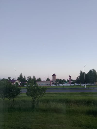The image depicts a semi-rural landscape with a road in the foreground and buildings in the background. The sky is clear with a crescent moon visible. A two-lane road runs horizontally across the mid-ground, bordered by a grassy median and a fence. On the other side of the road, several buildings are visible, including houses with pitched roofs and a longer, rectangular building with multiple windows. Two prominent towers with red roofs are also present in the distance. The foreground consists of a dense, green grassy area with two small trees. The overall scene suggests a late afternoon or early evening setting.