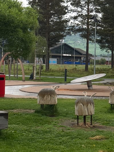 This outdoor scene captures a quiet recreational area or park, likely in Vinstra, Norway, under an overcast sky during daytime. The primary setting features a blend of green grass, various trees including tall conifers and smaller deciduous types, and a winding path constructed from wooden planks and gravel.
In the midground, several distinctive wooden structures are visible. These include multiple stout, log-like pieces with carved features resembling heads or faces, each fitted with ropes, potentially serving as seating or play elements. A prominent feature is a large, light-colored circular disc mounted on a dark cylindrical base, possibly an interactive spinning or balancing toy. A red trash receptacle is partially visible in the lower left.
The background is anchored by a contemporary building featuring a dark roof and extensive glass facades. Visible text on the building identifies "GROHE" and "SERVICE", while a large sign on its side clearly states "VINSTRA". Beyond the building, the landscape extends to reveal distant, rolling hills or mountains, characteristic of Norwegian geography. Several utility poles with lighting fixtures are also scattered across the background.
No individuals are present in the image, suggesting a tranquil moment in the park. The diffuse lighting indicates an overcast or cloudy day, casting soft shadows.