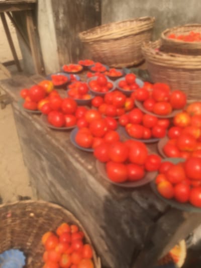 The image depicts a produce stall in Abagbo, Nigeria. Numerous bright red tomatoes are the primary subjects, presented in various quantities. Many individual servings of tomatoes are arranged on small, round, possibly blue or grey plates, clustered across a roughly textured, brown wooden table or stall surface. Several larger, woven baskets, appearing to be made of natural fibers, are visible in the background and foreground, some containing additional tomatoes. The background includes light-colored, possibly painted or stucco walls on the left and a darker, textured surface behind the upper baskets. No people are visible in the scene. The overall image is significantly blurred, making fine details indistinct.