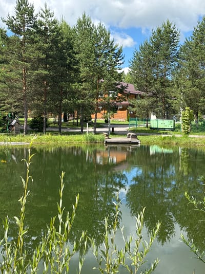 In Vorsma, Russia, a tranquil scene unfolds beside a body of water reflecting the sky and surrounding trees. A wooden house with a dark roof sits nestled among tall pine trees. In front of the house, a small wooden pier extends into the water, with two simple wooden benches on it. To the left of the pier, a miniature windmill structure and a green playground set are visible. A bright green generator unit is positioned behind a fence further down the bank, near a paved pathway. The water is a deep green, with lush vegetation growing along its edge and reflected in its surface. The sky above is a mix of blue and white clouds, casting subtle shadows on the landscape.