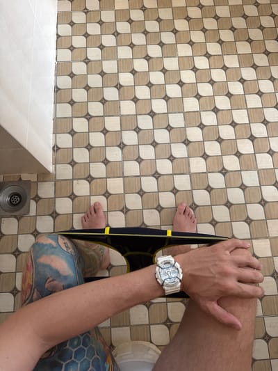 The image depicts a person sitting on a toilet in a bathroom. The person is holding a pair of black underwear with a yellow waistband. The person has fair skin, a white watch on their left wrist, and tattoos on their left leg. The floor is covered in light and dark brown patterned tiles. A drain is visible in the corner of the room. The exact location within Nam Nha Trang, Vietnam, cannot be determined from the given image.