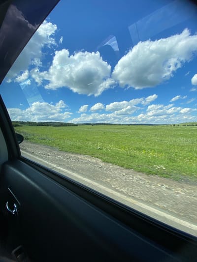 A view from inside a vehicle shows a rural landscape under a blue sky filled with white, fluffy clouds. The foreground is dominated by the dark interior of the car, including a door panel and a silver door handle. To the right, through the car window, a bright green, grassy field stretches to the horizon. A dirt road runs alongside the field. In the distance, a line of dark green trees marks the edge of the field. The overall scene depicts a sunny day in a rural area, possibly in the city of Yuldybayevo, Russia, as suggested. There are no people or animals visible in the frame.
