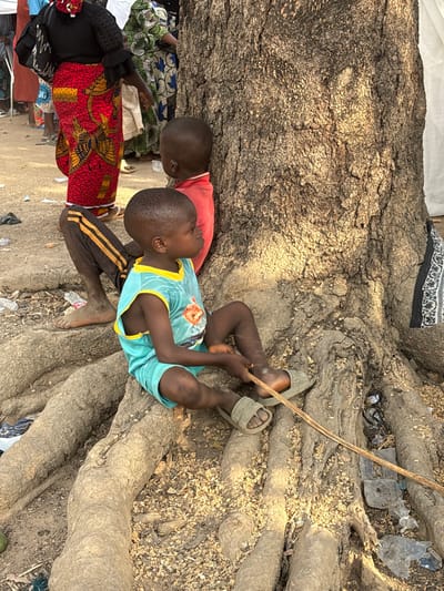 A large tree's roots surround two young children. One child lies against the trunk, wearing a red shirt and dark pants with gold stripes. The other child, in a teal tank top and sandals, sits among the roots, holding a thin stick. Adults in colorful patterned clothing stand nearby. Litter is scattered among the tree roots.