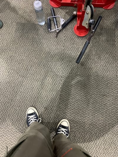 The image shows a pair of black canvas shoes with white soles, worn by an individual standing on patterned carpet. The person is wearing brown cargo pants and only their feet are visible in the image. The carpet is beige with a dark chevron pattern. In the top right corner, there is a piece of exercise equipment with red and silver components and two black handles. A clear plastic bottle containing liquid is lying on the carpet near the equipment. The location is in Kecamatan Kartasura, Indonesia. The coordinates are -7.567 latitude and 110.757 longitude. The scene is typical of a gym or fitness center.