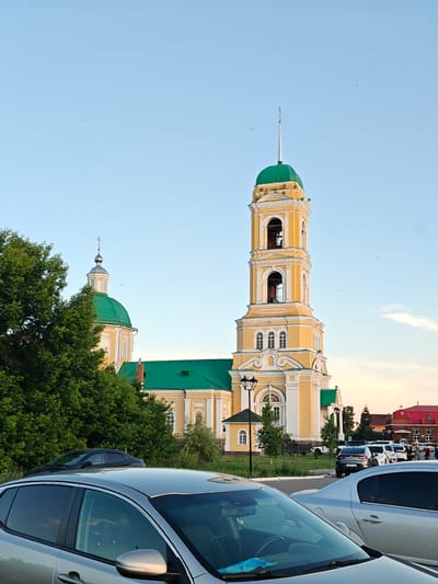 This image captures the Orthodox Christian Church of the Annunciation in Nikolo-Beryozovka, Russia, under a clear, light blue sky indicative of late afternoon or early evening. The prominent architectural complex features a main building and a tall bell tower, both primarily colored yellow with white architectural trim.
The multi-tiered bell tower, positioned centrally, is topped with a vibrant green, pointed spire culminating in a silver cross. Arched openings housing bells are visible in its upper sections, and minor vegetation growth is apparent on some cornices. To the left and slightly behind the bell tower, a larger, rounded dome, also green, is partially visible, topped with a smaller silver cupola and cross. Connecting sections of the church complex feature green roofs.
The foreground is occupied by parked vehicles, including the front section of a silver SUV on the left and a silver sedan to its right. Further back, a dark grey sedan is parked near a patch of green grass. A black ornate street lamp post stands in front of a small, yellow auxiliary building with a green roof, located to the right of the bell tower's base. Beyond this, a paved parking area contains several more cars, including white and dark-colored vehicles.
Lush green trees partially obscure the church structure on the left side of the frame and appear around the base of the buildings. In the distant background on the right, a cluster of buildings with red roofs and light-colored walls indicates a townscape. Several indistinct figures, likely people, are visible near these distant buildings, suggesting human presence and activity in the area. The sky, while mostly clear, shows a slight orange hue on the far right horizon, affirming the late hour. Small dark specks, possibly birds or insects, are scattered across the upper sky.