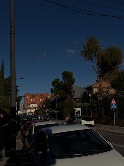 The image depicts a daytime street scene in Pozuelo de Alarcón, Spain, under a clear blue sky with sparse white clouds. The perspective is from street level, looking down a road lined with parked vehicles.
In the immediate foreground, the hood and windshield of a white car are visible, parked on the right side of the frame. To the far left, a tall, dark grey utility pole stands vertically, casting a long shadow towards the left. Overhead, multiple dark utility lines traverse the upper portion of the frame.
Along the left side of the street, a row of additional cars is parked, including red and dark-colored sedans or hatchbacks. Beyond the parked cars, dense green foliage, primarily trees and bushes, lines the road. A green rectangular street sign is partially visible amidst the foliage on the left. Further down the street on the left, a prominent multi-story building constructed of red brick with numerous small windows and a distinct roofline featuring dormer-like structures is visible. Below this, a white building is partially discernible.
On the right side of the street, a paved road surface is visible, bordered by a sidewalk or shoulder. A white Nissan Leaf electric car, identifiable by its rear design and Spanish license plate "2645 MHX", is parked on the right. Behind this vehicle, a light grey utility box or structure is present. A blue square sign with white pictograms indicating a pedestrian and bicycle crossing is mounted on a pole further down the right side of the street. The right side of the road is also heavily bordered by green foliage, primarily tall trees and dense bushes, behind which a brown-toned building with a dark pitched roof is partially visible.
No individuals are discernible in the scene. The lighting indicates direct sunlight, casting distinct shadows.
