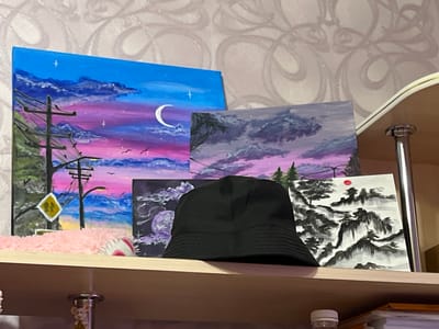 A shelf with several paintings and a black bucket hat is displayed against a wallpapered wall. The shelf is made of light-colored wood and is supported by metal fixtures.
The paintings vary in style and subject matter:
* A large canvas on the left depicts a vibrant sky with hues of blue, pink, and purple, featuring clouds, a crescent moon, birds, a utility pole with wires, and a yellow diamond-shaped sign with two figures.
* A smaller canvas to the right of the first one shows a purple and pink sky with dark clouds and silhouetted trees.
* Another painting, partially obscured by the hat, displays a dark, moody scene with a purple celestial body and swirling patterns.
* To the right, a painting rendered in a traditional East Asian ink wash style features a mountainous landscape with pine trees and a red sun.
A black bucket hat is placed prominently in the foreground, covering part of the paintings. Below the shelf, the edge of another shelf is visible with some books and a plastic water bottle. A decorative mouse figurine is also visible below the shelf. The wallpaper has a swirling pattern with a subtle shimmer. The scene is consistent with an indoor setting, possibly a home or studio. There is no information in the image to indicate a specific location in the city of Muravlenko, Russia.
