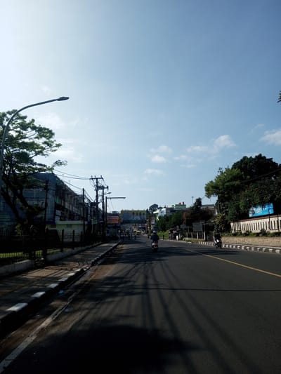A street scene photographed during daylight, in what may be a city or town. The sky is mostly clear, with patches of light cloud cover. A paved road occupies most of the lower frame, with strong shadows cast across its surface. A single yellow line marks the road's center. Along the left side of the road is a sidewalk and a low, black metal fence. Behind the fence are buildings, with one partly obscured by a tree. Utility poles with numerous wires run along the side of the road. On the right side of the road is a low wall, possibly a fence, with vegetation behind it. Two people riding motorbikes are visible on the road. The background shows more buildings and trees, suggesting a built-up area. No indication suggests that this image is from Nhot Chang, Laos.