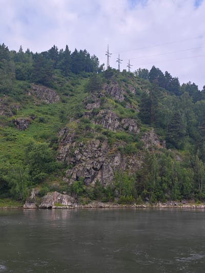 A wide, dark gray river flows in the foreground, with ripples and some white foam visible on its surface. On the right bank of the river is a steep, rocky hill covered in dense green vegetation and trees. The rocks are gray and jagged. At the top of the hill, several tall, thin power line towers with wires stretching between them are visible against a cloudy, pale sky. Some lighter green trees are scattered among the darker green foliage on the hillside. In the lower left corner of the image, on the bank of the river, there is a rocky outcrop with some graffiti or markings on it and a red and white striped marker pole.