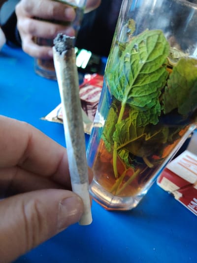 A hand holds a lit, rolled cigarette near a glass of amber liquid with green mint leaves. The cigarette's lit end is grey. The glass is tall and clear, possibly containing tea. Behind the glass and cigarette, blurred objects include a partially visible person holding a glass with a lighter-colored liquid, a pack with red and white design, and a gold foil. The background is a flat blue surface.