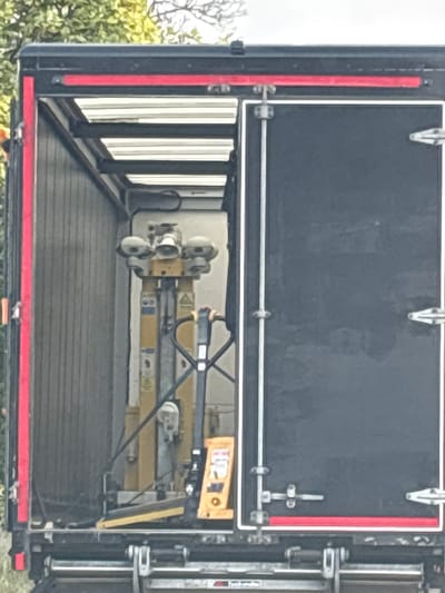 A black truck trailer is parked in South Staffordshire, United Kingdom. The rear doors of the trailer are open, revealing the interior. Inside the trailer, a yellow pallet jack is visible. The trailer is lined with grey vertical panels. A red stripe runs along the top edge of the trailer's opening. The background consists of green foliage.
