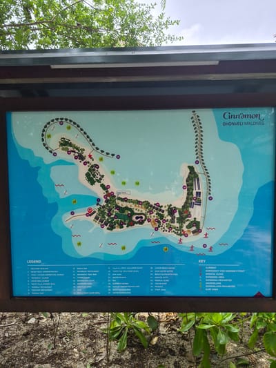 The image shows a map of the Cinnamon Dhonveli Maldives resort, located in Huraa, Maldives. The map details the layout of the resort, including bungalows, restaurants, pools, and recreational facilities. The key indicates locations of interest, such as the medical clinic, emergency fire assembly point, and swimming and snorkeling areas. The legend provides a list of key locations with their corresponding numbers. The image also contains a border and foliage.