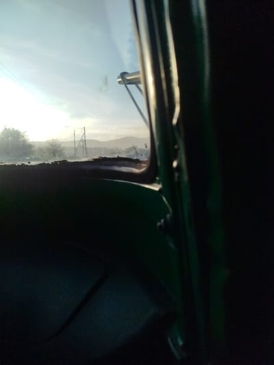 The image depicts a static scene captured from the interior of a vehicle, likely a green-painted automobile, looking outwards through a front window. The interior of the vehicle, including a dark green dashboard or seat in the lower left and a prominent dark green pillar on the right, is largely in shadow. A metallic component, possibly an antenna or mirror arm, is visible protruding from the vehicle's exterior into the upper right portion of the view.
Outside the vehicle, the environment is brightly illuminated under a clear, pale blue sky with diffused sunlight, suggesting either early morning or late afternoon. A flat, open landscape stretches towards a hazy horizon line marked by distant hills or mountains. In the midground, two utility poles with overhead lines are visible. Sparse trees and scrub vegetation are present on the left side of the frame, bordering what appears to be an unpaved road or dirt area. No individuals, animals, or specific actions are discernible within the frame. The scene is situated in Keekonyokie ward, Kenya.