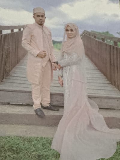A newlywed couple poses for a photograph on a wooden bridge in Babahjurong, Indonesia. The groom, wearing a pale peach-colored traditional outfit and a peached fez, stands on the left. The bride, in a matching pale peach-colored hijab and a white lace wedding dress, stands on the right. They are holding hands. The bridge is made of wooden planks and has a wooden railing. The background shows a cloudy sky and some greenery. The overall lighting suggests it might be an overcast day.