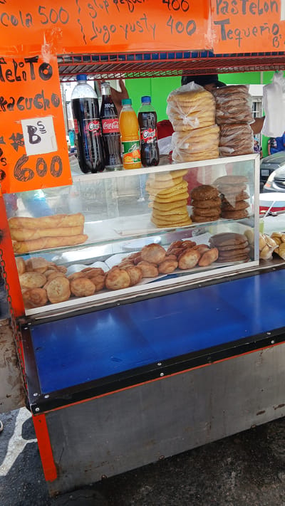 This is an outdoor food cart in Puerto La Cruz, Venezuela. The cart displays various breads, pastries, fried dough, and beverages. The cart has signage in Spanish advertising prices of items, including "cola" at 500, "glup" and "Jugo yusty" at 400, "pastelon" and "Tequeño" for an unspecified price, and "Felito Aragucho" at 600. The beverages visible on the cart are "Glup" and "Coca-Cola". A person is partially visible near the top-left edge of the image. The cart has a blue side panel. The ground near the cart appears to be concrete or asphalt.