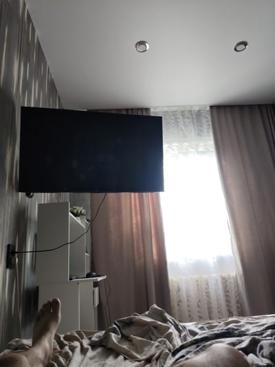 The image depicts an interior view of a bedroom. A large, black flat-screen television is mounted on a wall with striped wallpaper. Below the TV, there is a white shelving unit with some items on it, including what appears to be a potted plant with white flowers and a computer mouse. To the right of the shelving unit, a window is covered by sheer patterned curtains and heavier drapes. The room has a white ceiling with two circular recessed lights. In the foreground, a person's bare foot is visible, resting on a crumpled bed with a patterned duvet. The scene appears to be located in a bedroom, likely in Shimanovsk, Russia, given the context provided. The lighting suggests it is daytime, with bright light coming through the window.