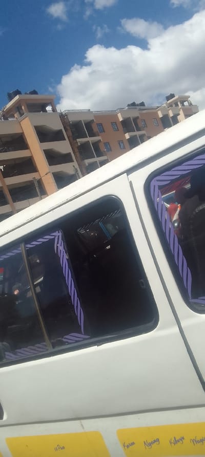 The image depicts a scene in the city of Naivasha, Kenya, captured from the interior of a white vehicle, likely a matatu. The perspective is upward and angled, showcasing a multi-story building against a partly cloudy blue sky.

The sky in the upper left quadrant is clear blue, transitioning to a dense cluster of white cumulus clouds across the upper right.

In the midground, a light peach and beige colored multi-story building stands, appearing to be an apartment or commercial structure. It features multiple floors with balconies or recessed windows. Several dark water tanks are visible on the rooftop. A thin, light-colored vertical pole is discernible in front of the building's left section.

The foreground is occupied by the white side panel of the vehicle. Two dark-tinted windows are prominently featured, each adorned with purple and white diagonally striped tape along its inner perimeter. Faint reflections or silhouettes of at least two occupants are visible within the windows; one individual in the right window wears a dark garment with a hint of red. Below the windows, on the lower white panel, several rectangular yellow stickers are affixed. Text visible on these stickers includes "11 Pass" on one, and the names "Karen," "Ngang," "Kibuyu," and "Wagia" on others, likely indicating route points or associated names. The vehicle itself shows no overt actions or interactions.