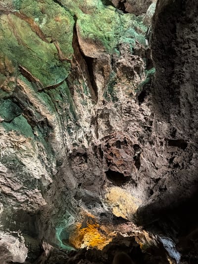 This image shows the interior of the Cueva de los Verdes, a lava tube located in Haría, Lanzarote, Spain. The walls and ceiling of the cave exhibit rugged, volcanic rock formations, illuminated with artificial lighting that casts green and yellow hues. The rock surfaces display variations in texture and color, ranging from dark gray to light green and brown. The lighting highlights the irregular shapes and crevices of the rock formations. There are no people visible.