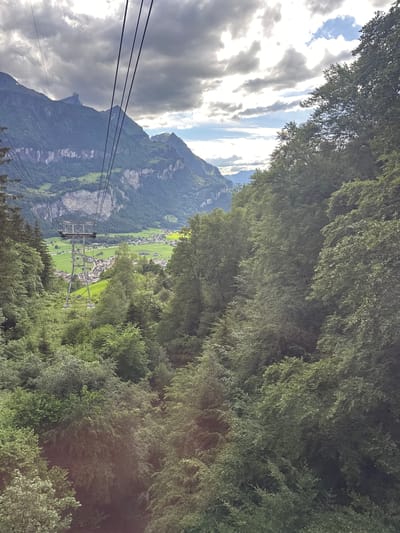 This is a medium-angle, exterior, eye-level shot taken from a cable car. The image captures a scenic mountainous landscape in the region of Geissholz, Switzerland. In the foreground, dense green trees line both sides of the frame, with the cable car wires stretching diagonally across the upper portion of the image. A steel cable car support tower is visible in the mid-ground, with a village nestled in the valley below. The valley floor is a patchwork of green fields and buildings, leading up to towering, rugged mountains in the background. The sky is partly cloudy with areas of bright sunlight breaking through the dark, dramatic clouds. No people are visible in the image.