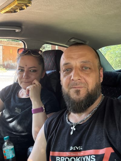 This is a selfie taken inside a vehicle, likely a car, with two people visible. The image is oriented sideways.

In the foreground, a woman is positioned towards the bottom left of the frame. She has her head resting on her hand, with sunglasses perched on top of her head. She is wearing a black t-shirt with a pattern of what appears to be the "Miu Miu" logo repeated. On her left wrist, she wears a watch with a purple band. Her expression is neutral, with her eyes looking towards the camera.

Behind and to the right of the woman is a man. He is facing the camera, with a beard and short hair. He is wearing a black t-shirt that has text on it, including "DEVIL" and "BROOKLYNS" with "Do Own Reuse" in between. He is also wearing a silver chain with a cross pendant. His expression is neutral, and he is looking at the camera.

The setting appears to be a car interior. The car's headliner is visible, as is a car door handle. Through the window, glimpses of greenery and what looks like a building with brick and orange siding can be seen, suggesting an outdoor environment. The lighting suggests it is daytime.

There is a gold-colored object to the left of the woman, possibly an ornament. A bottle of drink is visible on the floor of the car to the right of the woman.

The overall scene is a casual selfie taken by two individuals in a vehicle.