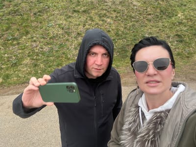 Two individuals are captured in a close-up, waist-up selfie-style photograph within Despotovac Municipality, Serbia. The scene depicts a male subject on the left, wearing a black hooded jacket with the hood raised and a black undershirt. He has light skin and a neutral expression, looking directly at the camera. His right hand, adorned with a silver-colored ring on the fourth finger, holds a green smartphone horizontally, angled towards the viewer, suggesting he is operating the device to capture the image. To his right stands a female subject with dark hair pulled back, wearing gold-framed cat-eye sunglasses with dark lenses, a white hooded sweatshirt, and an olive-green puffer jacket featuring a prominent multi-toned faux fur collar. She displays gold-colored hoop earrings and a slight pink blush on her cheeks, with her mouth slightly open, also looking towards the camera. The background consists of a gently sloped, partially green and partially dry grassy area with scattered broad-leafed plants. The immediate foreground shows a light grey paved surface, appearing somewhat damp. The lighting is diffuse, indicative of an overcast day.