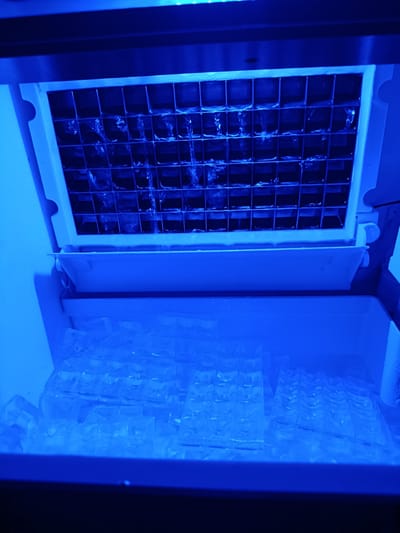 The image shows an ice maker with ice cubes in a bin and ice being formed in a grid. The scene is lit with blue light. There are no people present. The image does not provide any information about the location in the city of Pishchane, Ukraine.