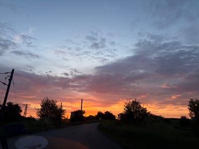 The image depicts a road leading into the distance, framed by trees and vegetation on either side. In the background, there is a power plant with a tall chimney. The sky is filled with clouds, with colors ranging from blue to pink and orange, indicating either a sunrise or sunset. Power lines and poles are visible on the left side of the road. The image was taken from inside a car. The location is Obruchishte, Bulgaria.
