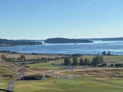This is a daytime, wide-angle, eye-level shot of Chambers Bay Golf Course in University Place, Washington, United States of America. The golf course occupies the foreground, with rolling green fairways and brown, dry roughs. A winding asphalt path is visible, with a golf cart traveling on it. A building with a green, grass-covered roof sits near the path. In the middle ground, a parking lot is filled with cars, and a few trees are scattered across the landscape. Beyond the golf course, a body of water, likely Puget Sound, stretches to the horizon. A large, tree-covered island is situated in the water, with a ferryboat visible near its shore. The distant background shows a hazy coastline with hills. The sky is clear and blue.
