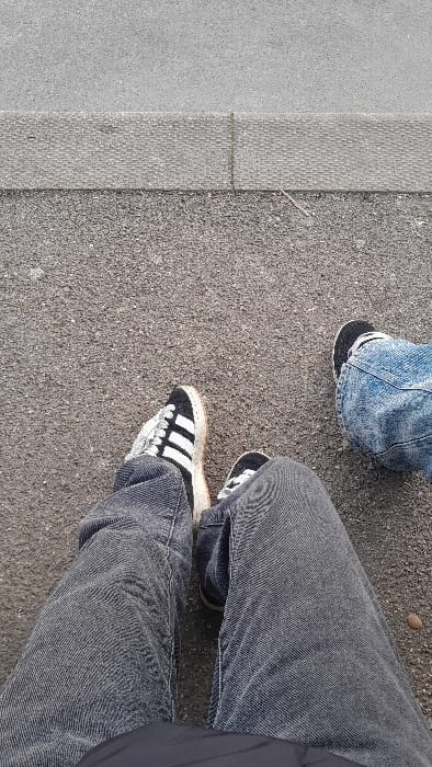 A top-down forensic image depicts the lower extremities of two individuals on an asphalt surface in Souffelweyersheim, France. The individual on the left wears faded dark grey jeans and black and white Adidas sneakers with white laces and tan-colored soles. The individual on the right wears distressed light blue jeans and a dark sneaker with a visible white stripe. Both subjects' feet are positioned on a dark grey, textured asphalt ground. A light grey concrete curb with a segmented, textured surface runs horizontally across the upper frame, separating the asphalt from a lighter, smoother paved area. A small, light-colored stick lies on the asphalt near the curb, and a pebble is visible near the right individual's foot.
