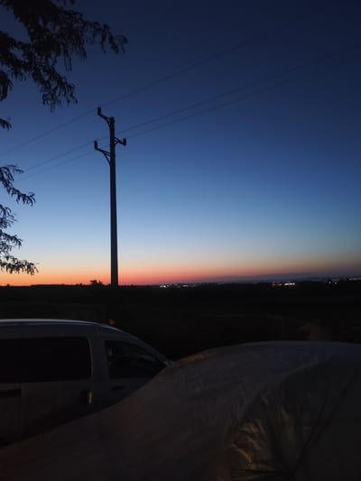 This image captures a serene twilight scene in Ravadinovo, Bulgaria. A dark tree branch extends from the upper left, its silhouette stark against a gradient sky that transitions from deep blue at the top to shades of orange and pink near the horizon. A single utility pole stands vertically in the center, supporting several power lines that stretch across the frame. In the foreground, the top of a silver van is visible on the left, its windows dark. To the right, a large object covered with a textured, dark material occupies the lower portion of the image. The distant horizon is a dark silhouette of land, punctuated by faint lights suggesting a settlement or town. The overall atmosphere is tranquil, with the fading light creating a peaceful ambiance.