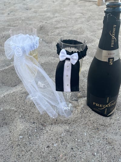 A bottle of Freixenet Cordon Negro Brut sparkling wine rests on a sandy beach, alongside two champagne flutes. The scene suggests a celebration, possibly a wedding or anniversary, given the festive attire on the glasses. One flute is adorned with a black band featuring a white bow and three small rhinestones, resembling a tuxedo. The other flute is decorated with delicate white lace, pearls, and a fabric rose, evoking a bridal veil. The champagne bottle, a classic black bottle with gold lettering, is positioned above the flutes, also lying on the sand. The sand itself appears to be a light beige color, with small shells and pebbles scattered across its surface, typical of a beach environment. The lighting suggests it is daytime, with no strong shadows indicating harsh midday sun. The overall impression is one of a romantic or celebratory occasion taking place in a relaxed, beach setting. The visible text on the bottle reads "FREIXENET", "SECO", "GRAN SELECCION", "NEGRO", and "CORDON".