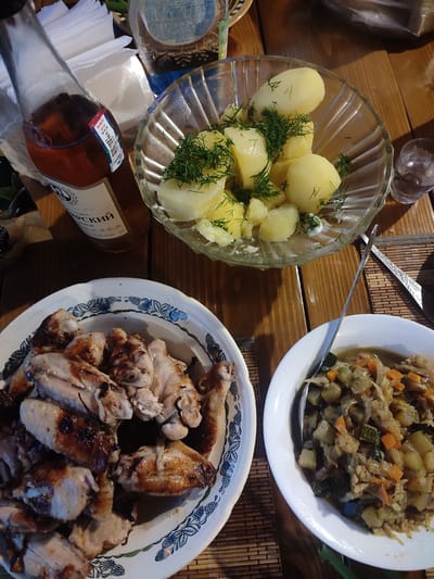 The image shows a table setting with several dishes and a bottle of liquid, likely a meal in Il'bukhtino, Russia. In the foreground, a white bowl contains what appears to be roasted chicken wings, some with visible char marks. To the right, another white bowl holds a mixture of chopped vegetables, possibly stewed zucchini, carrots, and onions. In the upper portion of the image, a glass bowl is filled with boiled potatoes, garnished with fresh dill. A bottle with a pinkish liquid and a label is positioned to the left of the potato bowl, and a small glass containing a clear liquid is to the right. The table is made of wood, and a woven placemat is visible beneath some of the dishes. The background is slightly blurred, but a stack of white napkins can be seen behind the bottle. The overall scene suggests a home-cooked meal or a casual dining experience.