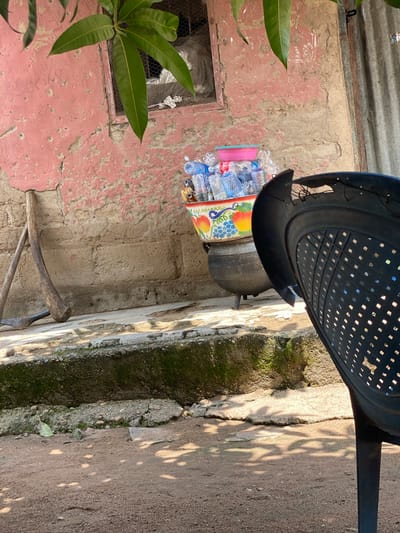 The image depicts an outdoor scene, likely in a residential area of Daniel, Nigeria. In the foreground, a dark plastic chair is partially visible, facing away from the viewer and angled to the right. Behind the chair, a collection of plastic water bottles is gathered in a large, decorative bowl. This bowl, featuring images of fruits like apples and grapes, sits atop a black, cast-iron pot with three legs. The pot is positioned on a raised concrete ledge, which also holds two wooden farming tools, leaning against a textured, pinkish-red wall. The wall is weathered and shows signs of cracking and peeling paint. To the right of the wall, a section of corrugated metal siding is visible. The ground in front of the ledge is made of reddish-brown dirt, with dappled sunlight creating shadows. Overhanging the scene from the top left are several large, green mango leaves. The overall impression is one of a functional, perhaps somewhat rustic, outdoor living or workspace.