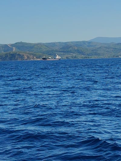 A wide shot captures a calm blue sea under a clear sky with distant green hills. In the middle ground, a large cargo ship is visible near the coastline of Paleokastro, Greece. The ship appears to be anchored or moving slowly. The water is rippled, suggesting a gentle breeze. The landscape features rolling hills covered in vegetation, with a few dirt paths winding up their slopes. The overall scene is serene and depicts a typical coastal view.