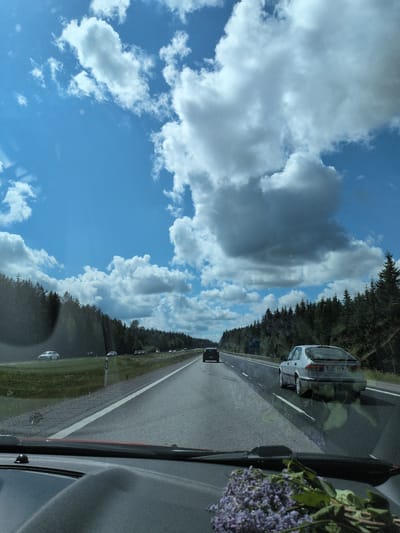 The image captures a forward-facing view from inside a vehicle on a multi-lane highway in the Heinola region of Finland, observed on a bright, partly cloudy day. The sky presents a blend of clear blue areas and large, white cumulus clouds, with some clouds casting shadows.

The highway features at least two lanes of travel in the observer's direction, separated by white solid and dashed lines, and is flanked by a grassy median strip and dense coniferous forests on both sides. On the left side of the road, a Finnish road sign is visible, displaying text that includes "LINJA PÄÄTTYY" (Line Ends) and "PEKALA".

Traffic is present, moving in the same direction. Immediately ahead in the right lane is a silver three-door Saab 9-3 hatchback (first generation, likely 1998-2002 model), identifiable by its rear profile and the Finnish license plate "AZT-869". Further ahead in the left lane, a dark-colored sedan is visible. On the opposite side of the divided highway, a white sedan is visible on the grassy median or shoulder, with additional vehicles in the distant opposing lanes.

The foreground of the image shows the top portion of a vehicle's dark dashboard, including the base of the windshield and a wiper blade. A bouquet of purple flowers, consistent with lilacs, is placed on the dashboard in the lower right section of the frame. Reflections from the vehicle's interior are visible on the windshield. No individuals are clearly discernible within the scene.