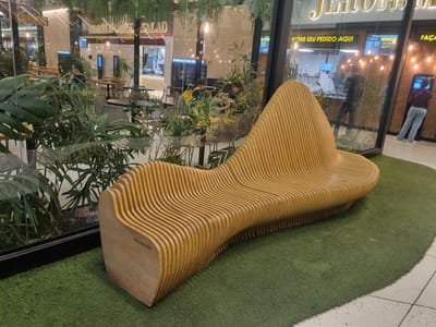 The image depicts a modern seating area, possibly within a mall or public space, featuring a distinctive, wave-like wooden bench. The bench is made of layered, curved wooden slats, creating an organic and sculptural form. It is positioned on a patch of artificial grass, which contrasts with the tiled flooring visible at the bottom right of the frame.

Through a large glass partition, a bustling indoor environment is visible. This area appears to be a food court or restaurant section. Signs with Portuguese text are present, including "INDROGRIND SALAD" and "RETIRE SEU PEDIDO AQUI" (Pick up your order here), suggesting a food service establishment. People are seen in the background, some standing at counters, others seated at tables. The lighting in the background is a mix of ambient and task lighting, with chandeliers and illuminated signs.

Specific people visible include a woman in blue jeans and a maroon top standing at a kiosk or ordering point, and a person in dark clothing and jeans in the food preparation area. Other individuals are partially obscured by plants and the glass. A young boy in a blue shirt is also visible sitting at a table with adults in the reflection.

The foreground is dominated by the unique wooden bench and the artificial turf. Lush green plants are strategically placed, adding a natural element to the modern design and partially obscuring the interior view. The overall impression is one of a contemporary, well-designed public space that blends natural elements with modern architecture and offers a comfortable seating option. The location is identified as Araçariguama, Brazil.