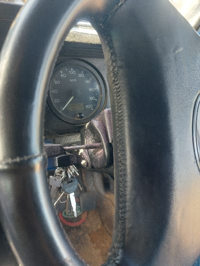 The image displays the interior of a vehicle, focusing on the driver's perspective from behind the steering wheel in the city of Kimamba, Tanzania. A black, leather-wrapped steering wheel occupies the foreground, partially obscuring the dashboard. Through the steering wheel's upper left opening, a circular speedometer is visible, featuring a black face marked in increments of 20 km/h, ranging from 0 to 200 km/h. The speedometer needle rests at 0 km/h, indicating the vehicle is stationary. Below the speedometer, a small unlit digital display is present, along with a single indicator light. To the right of the speedometer, a key ring with multiple keys is inserted into the ignition. The keys include a main silver-colored vehicle key with a black plastic head, several smaller metal keys, and a circular key fob with red and green elements. The dashboard surface surrounding the instruments and ignition appears dark and shows signs of dust or light debris accumulation. The vehicle's floor or footwell, visible beneath the ignition, has a worn, brownish texture.