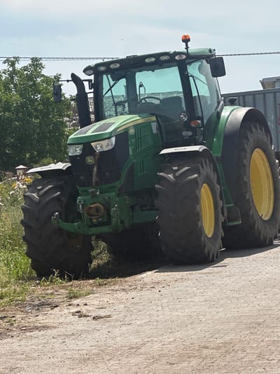 A green John Deere tractor is parked on a dirt road in Ostrówek, Poland. The tractor is facing the camera and is the main subject of the image. It has large, black tires with yellow rims. The tractor has a large cab with tinted windows and several lights on top. A black exhaust pipe is visible on the left side of the tractor. In the background, there are trees, power lines, and a grey trailer. The ground is covered in dirt and gravel, with some green vegetation on the left side of the image. The sky is cloudy.