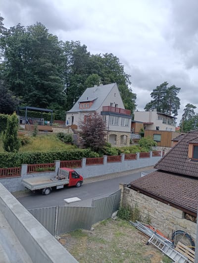 In Doksy, Czechia, a scene unfolds on a cloudy day with a focus on residential buildings and a red flatbed truck parked on a paved road. The truck, loaded with equipment including what appears to be a generator and tools, is positioned on the right side of the road. Adjacent to the road, a gray concrete wall topped with a wooden fence separates the street from a tiered landscape of greenery and a multi-story house.
The house, painted white with a gray roof, features multiple windows, balconies with red railings, and stone accents on its lower level. To the right of this house, another building with a wooden facade and a modern design is visible, with a ladder leaning against its side. In the foreground, a partial view of another building with a tiled roof and stone wall suggests ongoing construction or renovation, with materials like metal poles, pipes, and a ladder scattered on the ground. The background is dominated by lush green trees, characteristic of the surrounding natural environment.