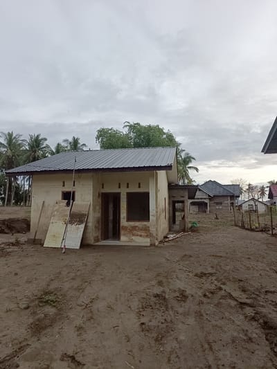 The image depicts a slightly overcast day in Meunasah Raya, Indonesia, with a focus on a partially constructed or renovated dwelling. The structure is simple, with cream-colored walls and a corrugated metal roof. To the left of the main entrance, two large, weathered boards are propped against the wall. A long pole leans against the house near the center. The ground in front of the house is unpaved and appears muddy, with tire tracks visible in places. In the background, several other houses with similar roofing are visible, along with palm trees characteristic of the tropical climate. A wooden fence with some vegetation behind it runs along the right side of the image. The overall scene suggests a rural or semi-rural setting undergoing some form of development or repair. There are no people or animals present in the image.