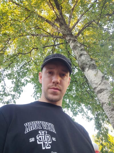 A low-angle, wide shot captures a man wearing a black baseball cap and a black t-shirt with white text. He is looking directly at the camera, with a neutral expression. The man is positioned in the foreground, with his upper body filling the bottom half of the frame. Behind him, a large birch tree with distinctive white bark and green leaves dominates the upper portion of the image. The sun filters through the leaves, creating a dappled light effect. The scene appears to be outdoors, under a canopy of trees, suggesting a park or natural setting. The lighting indicates it is daytime, likely late afternoon or early morning due to the warm tones. The text on the t-shirt is in a stylized font and reads "NEW YORK" in an arched pattern above a graphic logo and smaller text, though the smaller text is partially obscured and difficult to fully decipher. The image was taken in Polatsk, Belarus.