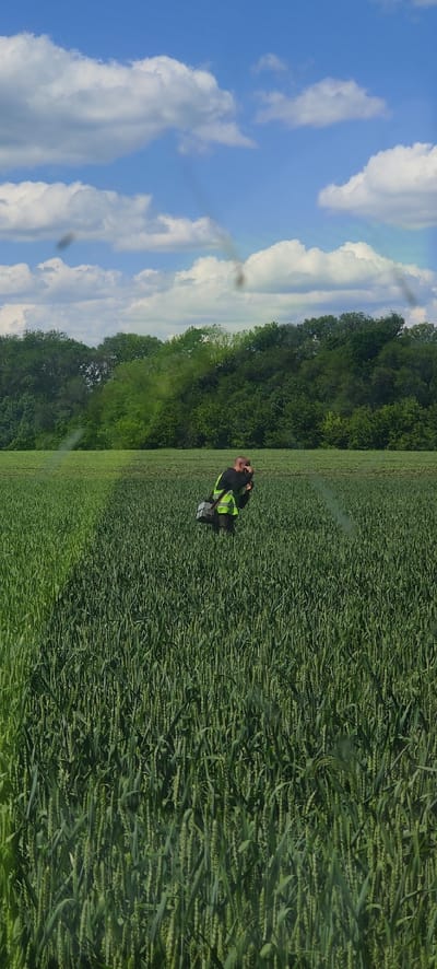 An individual is depicted squatting or kneeling in a large, lush green field under a bright blue sky with scattered white cumulus clouds. The person appears to be an adult male, wearing a dark top and a high-visibility yellow-green safety vest. He has a bald or very short-haired head. He is interacting with a piece of equipment, possibly a device for measurement or analysis, which is dark-colored with some metallic elements and rests on the ground in front of him.

The field is densely cultivated with green crops, likely a type of grain such as wheat, with visible developing heads. These crops fill the foreground and middle ground of the scene. In the background, a thick, dark green treeline or forest borders the field, extending horizontally across the image. A faint, light-colored plume, possibly smoke or vapor, is visible in the air behind the individual, though its origin is not clear.

The scene suggests agricultural work, such as crop monitoring, field inspection, or the use of specialized equipment in a rural setting. The time of day is bright daylight, likely midday or afternoon, under clear weather conditions. The location is an agricultural area in Pashchenivka, Ukraine. No visible text is present in the image.