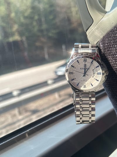 A metallic silver wrist watch is positioned vertically on the right side of the frame, resting against a dark, textured fabric surface and a lighter grey plastic vehicle interior trim. The watch features a white face with Roman numerals at the XII, IX, VI, and III positions, and a 24-hour sub-dial. The brand "ICEDOUT" is visible below the XII, with "TOP 1%" and "EXCLUSIVE QUALITY" inscribed below it. A date wheel displays the number '3' prominently. The watch has a multi-link metallic bracelet. The background, viewed through a window, is out of focus and shows a multi-lane asphalt road with blurred lines. A light-colored vehicle is partially visible on the road in the mid-ground. Dense green foliage and trees line the road further in the background. The scene is illuminated by natural daylight, with a bright glare reflecting off the watch's upper bezel. The overall perspective is from inside a vehicle traveling near Gorna Vasilitsa, Bulgaria.