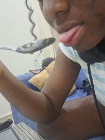 The scene is an interior setting in Close, Nigeria, depicting a partial view of an individual with dark skin. The person is seated or reclining on a blue upholstered sofa. Their right arm is bent, holding a silver-colored spoon containing a dark, viscous substance, which is positioned directly in front of their face. The person's tongue is extended from their mouth towards the spoon. They are wearing a light-colored, horizontally striped shirt, and black headphones are visible around their neck. A single, dark, curly strand of hair hangs near the person's face on the left. In the background, on the sofa, a woven, light-brown bag or pillow is visible, along with a clear plastic water bottle. The wall behind is plain and light-colored. On the floor to the lower left, a pair of black slip-on shoes are present, next to a surface with a striped, light-colored pattern.