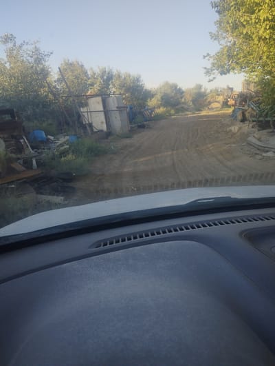The image depicts a view from inside a vehicle, looking out at a junkyard or scrap metal storage area. The foreground shows the vehicle's dashboard, with its textured surface and air vents. The windshield reveals a dusty, dirt road leading into the distance, with tire tracks visible. On either side of the road, there are piles of scrap metal, rusty machinery, and blue barrels. In the background, there are several structures, possibly sheds or makeshift shelters, along with trees and vegetation. The sky is clear and blue, suggesting daytime. The scene appears to be located in an outdoor industrial or rural setting. Without further information or context, it is not possible to determine the exact location in Margabat, Uzbekistan, or to identify specific people, actions, or interactions.
