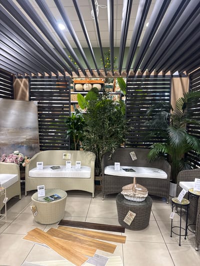 This appears to be a showroom display of outdoor furniture, potentially inside a store. The scene includes at least two sets of wicker-style patio furniture with white cushions. There are also round wicker tables, plants (including a palm), and what appears to be a piece of artwork displayed. Samples of flooring materials are arranged on the floor.
Given the presence of pricing tags and display materials, it is located in a retail or showroom environment, specifically in Pomezia, Italy.