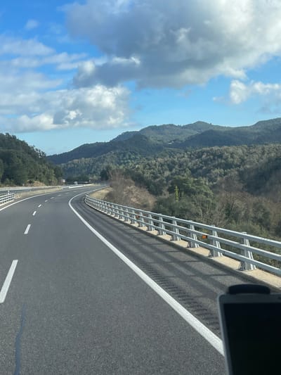 The image displays a two-lane asphalt highway curving left through a mountainous, forested landscape near Arbúcies, Spain. The sky is partly cloudy with blue patches and scattered cumulus clouds. The road features white solid and dashed lane markings. A silver-colored metal guardrail with multiple horizontal bars and orange reflectors lines the right side of the road, casting prominent shadows onto a concrete barrier and the road surface. Beyond the guardrail, a downward sloping, vegetated embankment transitions into a series of hills densely covered with a mix of evergreen and deciduous trees. Two light-colored vehicles are visible in the distance on the highway. A dark, rectangular object, consistent with a vehicle's dashboard component or a mounted device, occupies the lower right foreground. No individuals are present.